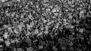 Melns un balts foto no liela cilvēku pūļa, kas piedalās protesta gājienā ar plakātiem.