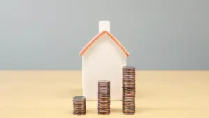 Mazs mājas modelis uz galda blakus augošām monētu kaudzītēm finanšu kontekstā