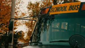 Dzeltena skolas autobusa priekšpuse ar atspulgu vējstiklā un rudens koku lapām fonā.
