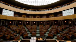 Tukša Saeimas plenārsēžu zāle ar koka galdiem un krēsliem politiskajām diskusijām.
