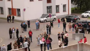 Cilvēku grupa pulcējas bruģētā laukumā pie daudzdzīvokļu ēkas, daži no viņiem tur papīra lapas.