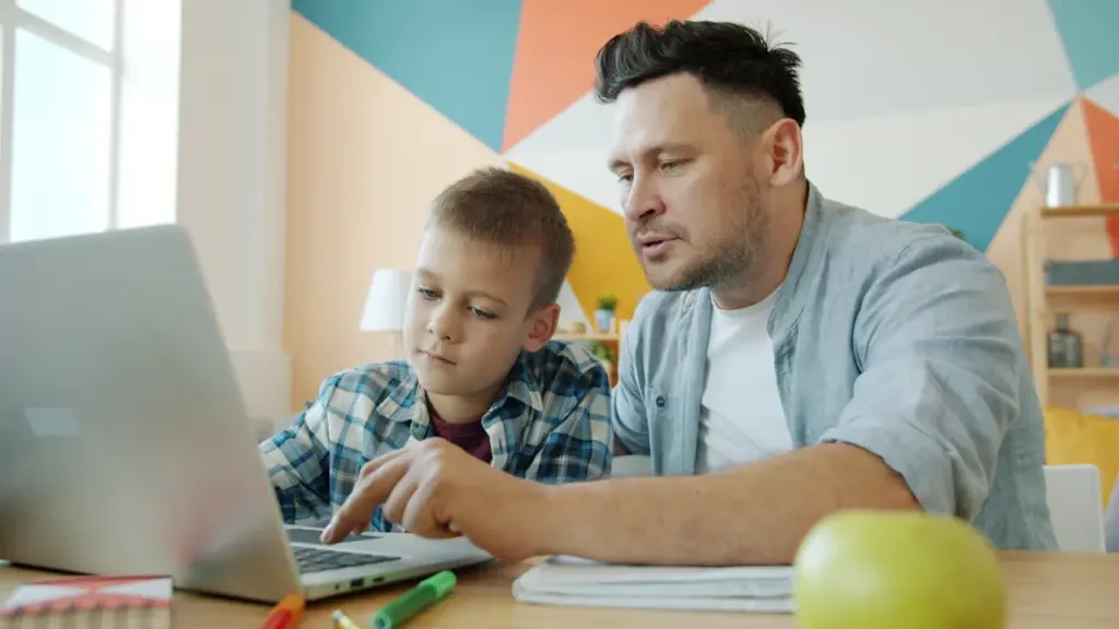 Tēvs palīdz dēlam ar mājasdarbiem pie datora modernā un mājīgā istabā.