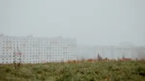 Balta daudzstāvu ēka Rīgā biezas miglas ieskautā, drēgnā oktobra dienā.