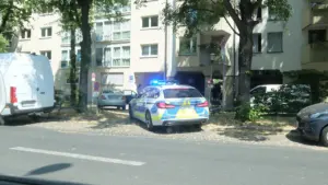 Policijas automašīna ar ieslēgtām bākugunīm stāv ielas malā pilsētvidē.